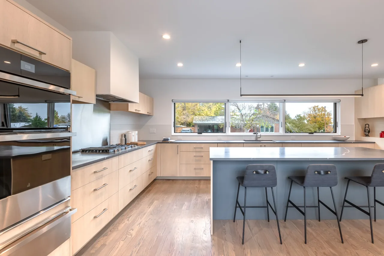 Contemporary kitchen features light wood cabinetry, a large horizontal window, and a pale blue kitchen island with seating.