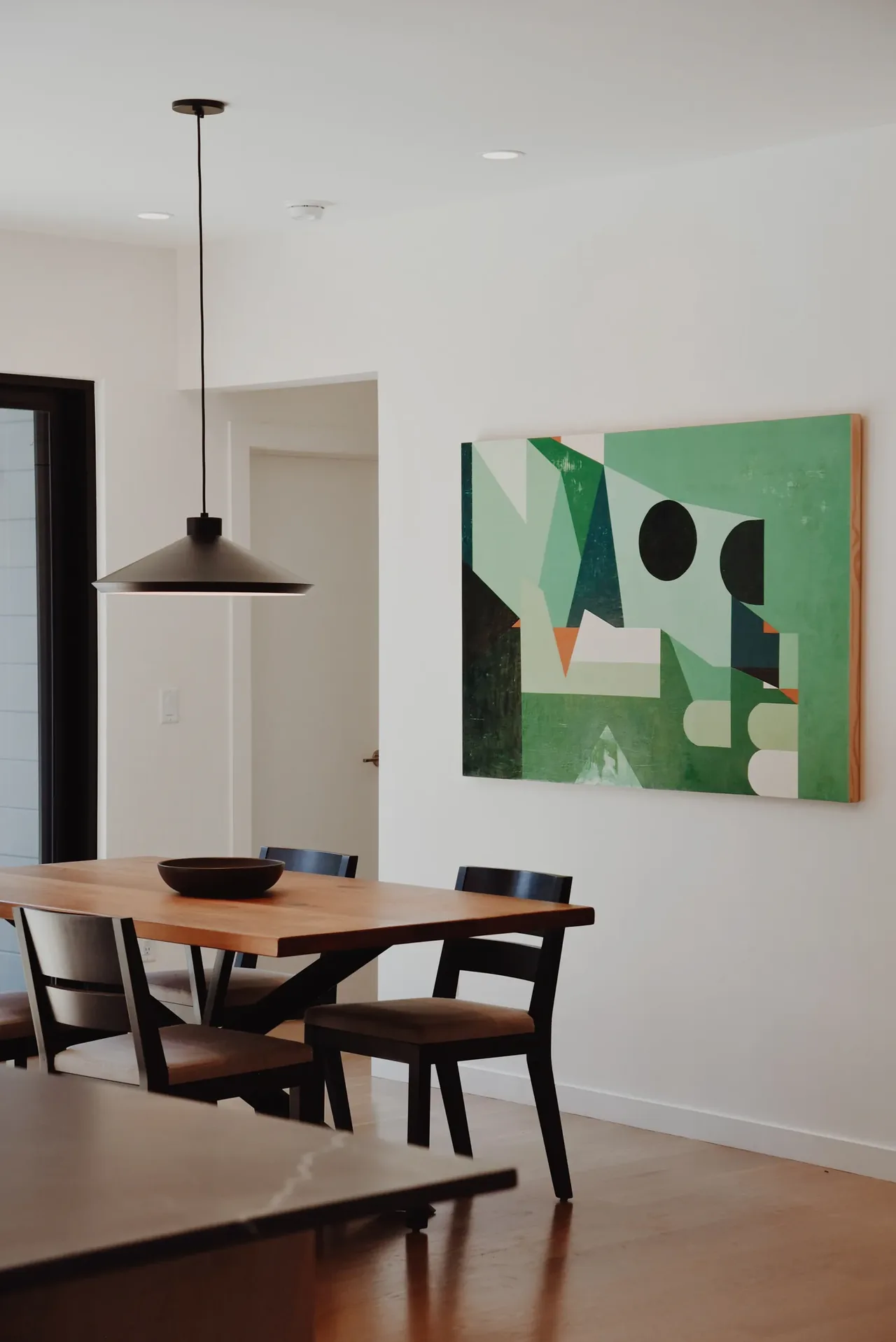 Dining area with a wood table, black chairs, and an abstract green painting on a white wall.