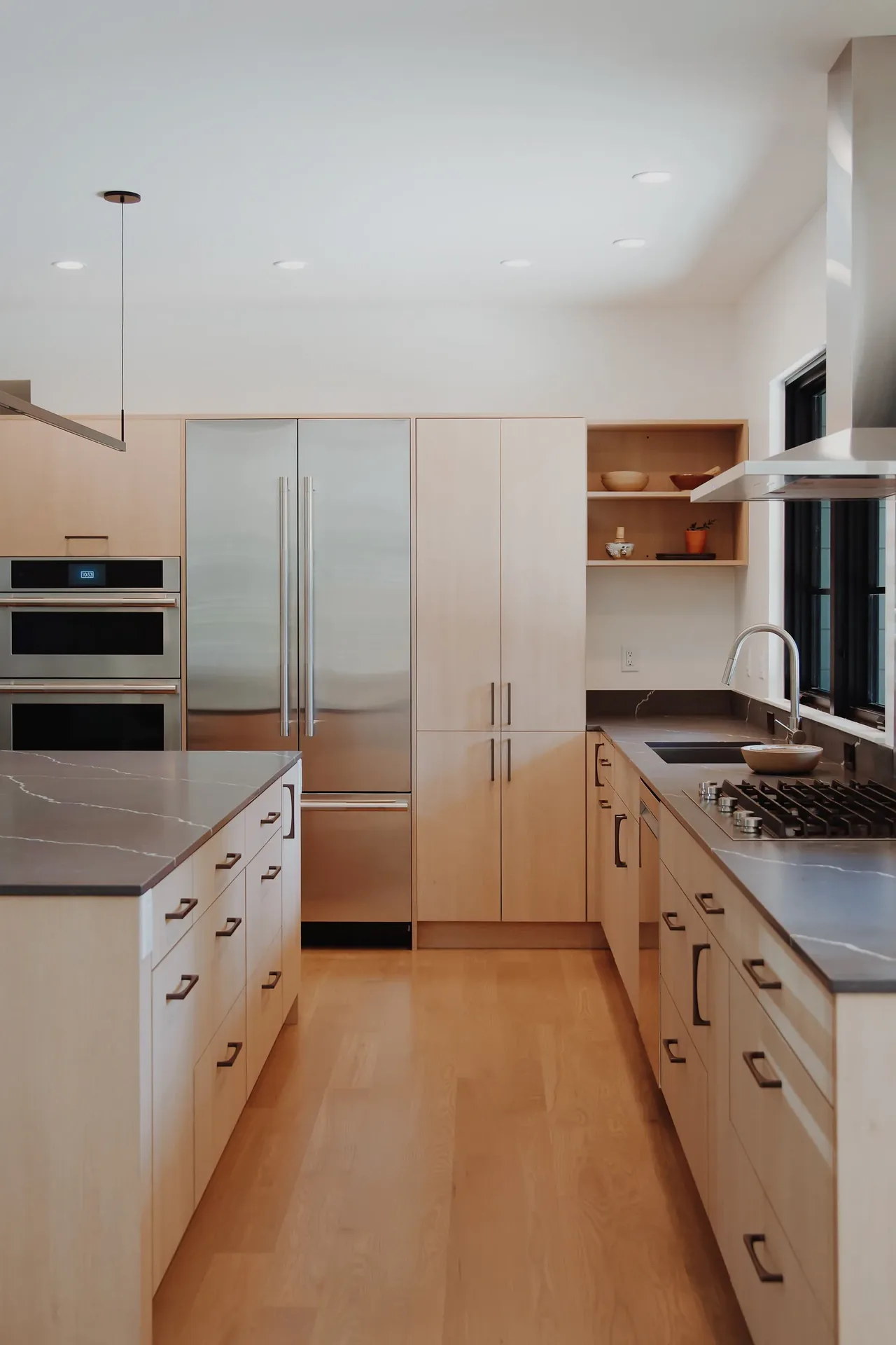 Light wood kitchen with integrated stainless steel appliances and a gas cooktop by a window.