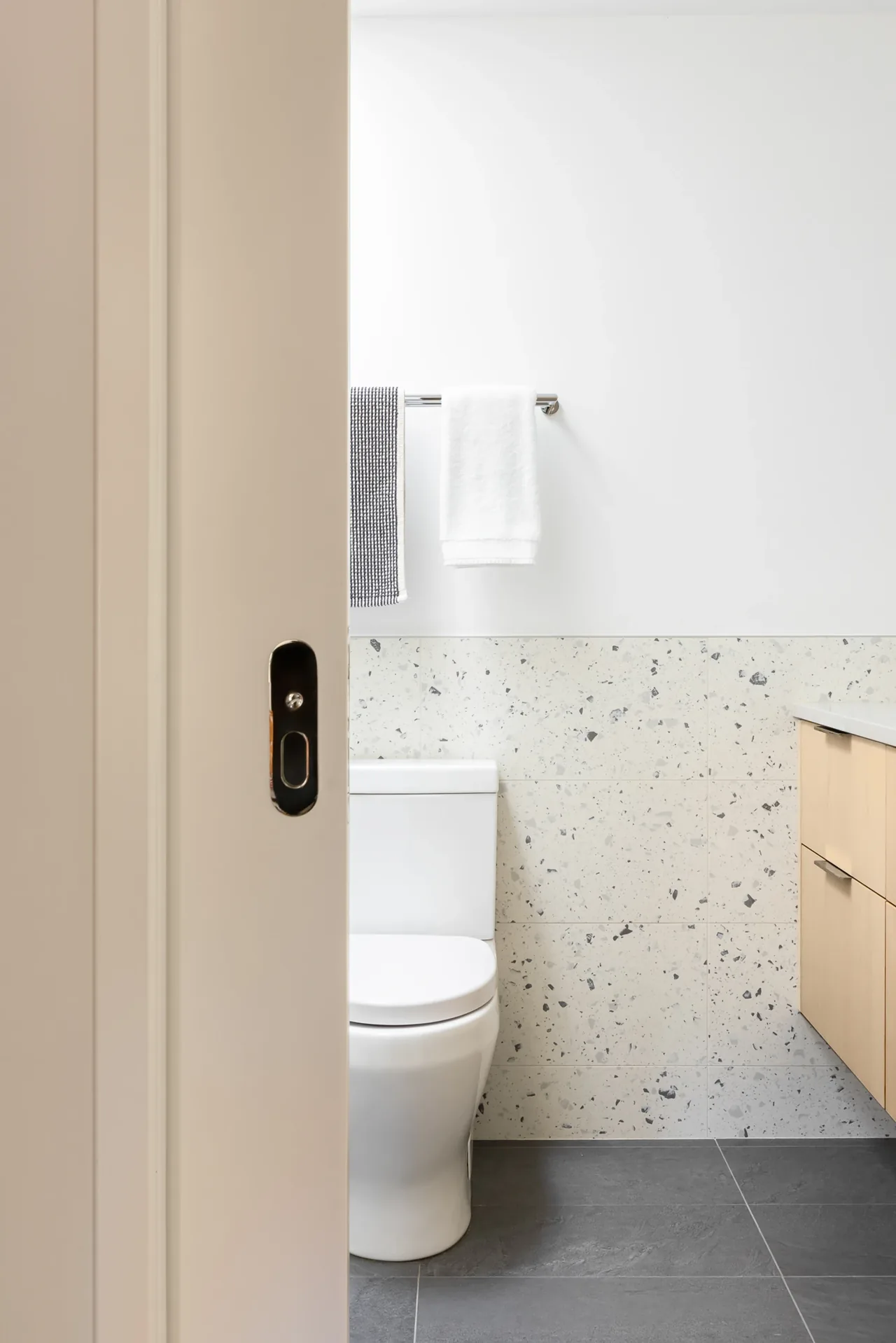 View into a bathroom with terrazzo walls, gray tile floor, and light wood vanity with white countertop.
