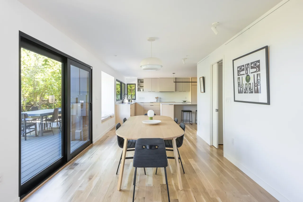 Dining area with wooden table and padded chairs adjacent to a kitchen, featuring light hardwood floors and large sliding glass doors.
