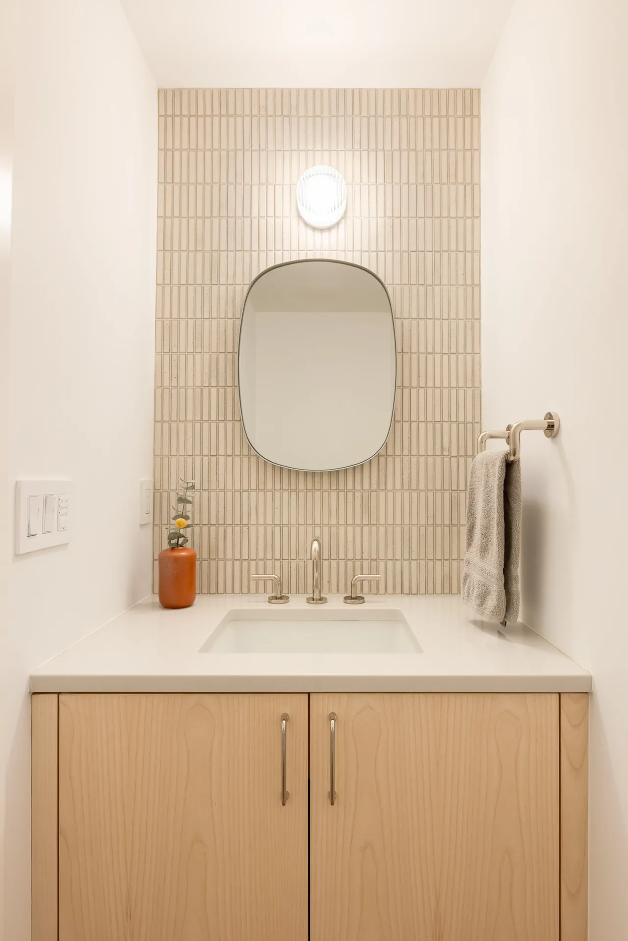 Minimalist bathroom vanity features light wood cabinetry, white countertop with undermount sink, vertical tile backsplash, and oval mirror under sconce light.