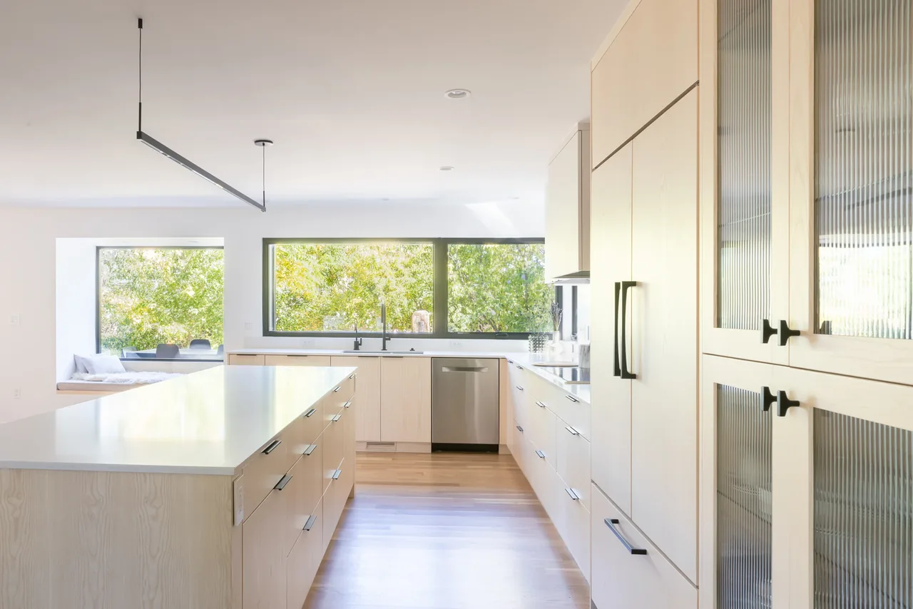 Kitchen featuring a large island, light wood cabinets with black handles, a panel-ready refrigerator, and floor-to-ceiling glazing overlooking greenery.