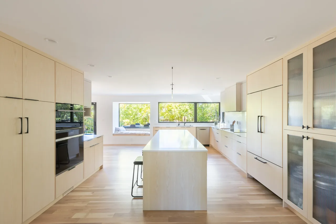Interior view of a kitchen featuring a large island, light wood cabinets, and floor-to-ceiling glazing for natural light.