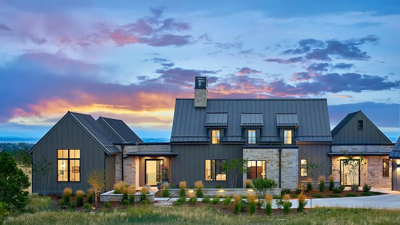 Exterior front view of a contemporary farmhouse at dusk with dark metal roofing and light stone walls under a colorful sky.