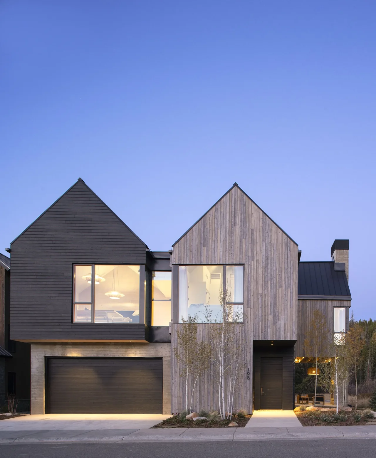 Modern two-story house with contrasting vertical wood siding and dark horizontal siding, large windows, and a concrete garage base under gable roofs.