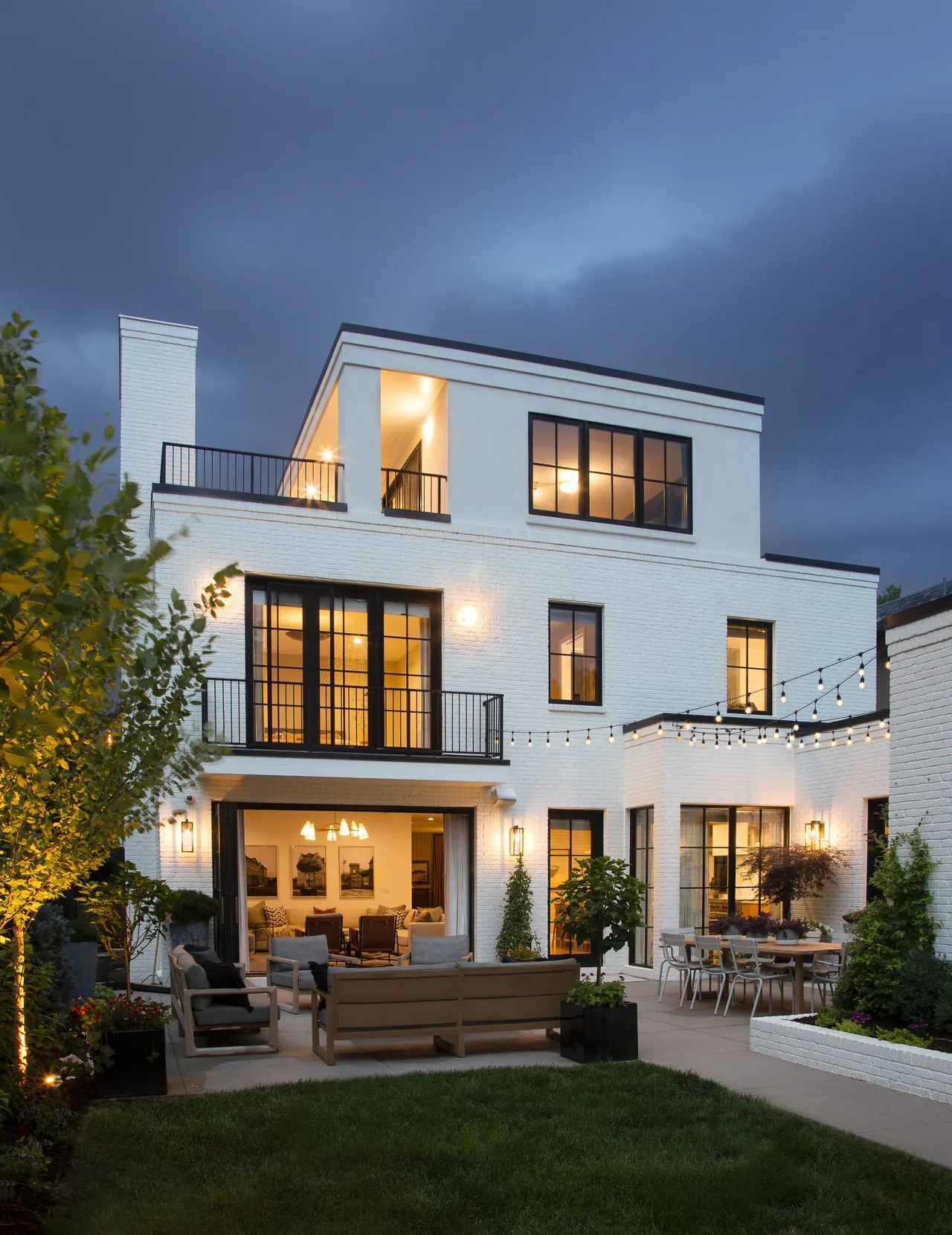 Three-story white brick house with black-framed windows, outdoor seating, dining area, and string lights illuminating the landscaped backyard.