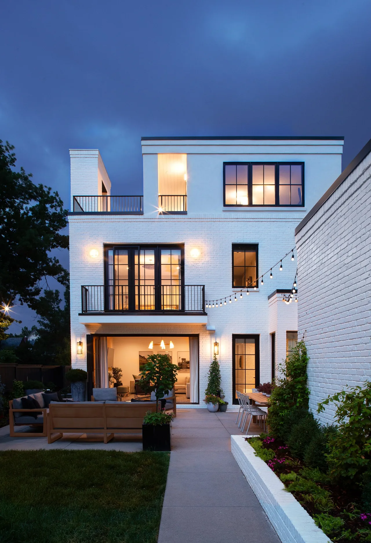 Rear exterior of Denver Residence 12 by Alvarez Morris, showing white brick, black window frames, and landscaped patio with outdoor seating.