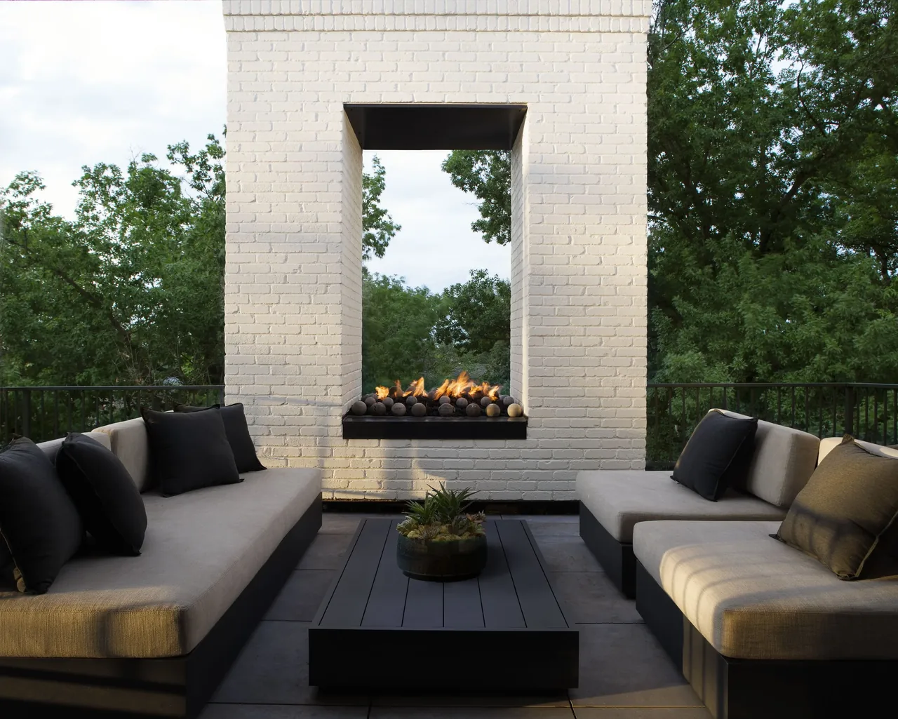 Modern outdoor patio with cream upholstered sofas, black pillows, and a white brick fireplace with fire.