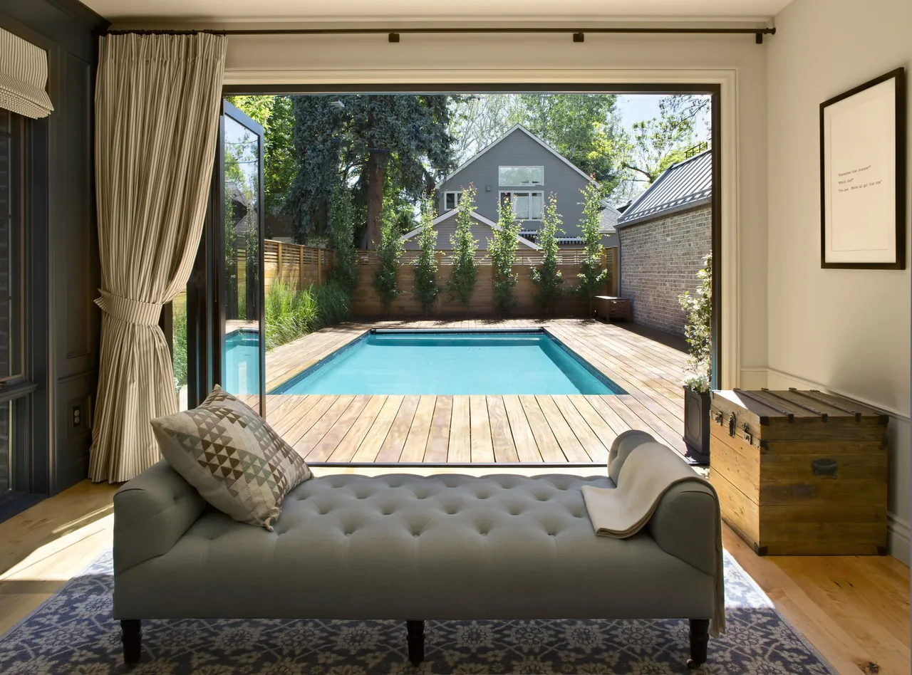 Interior room looking out through open glass doors to a wood deck and rectangular pool with gable-roofed house beyond.