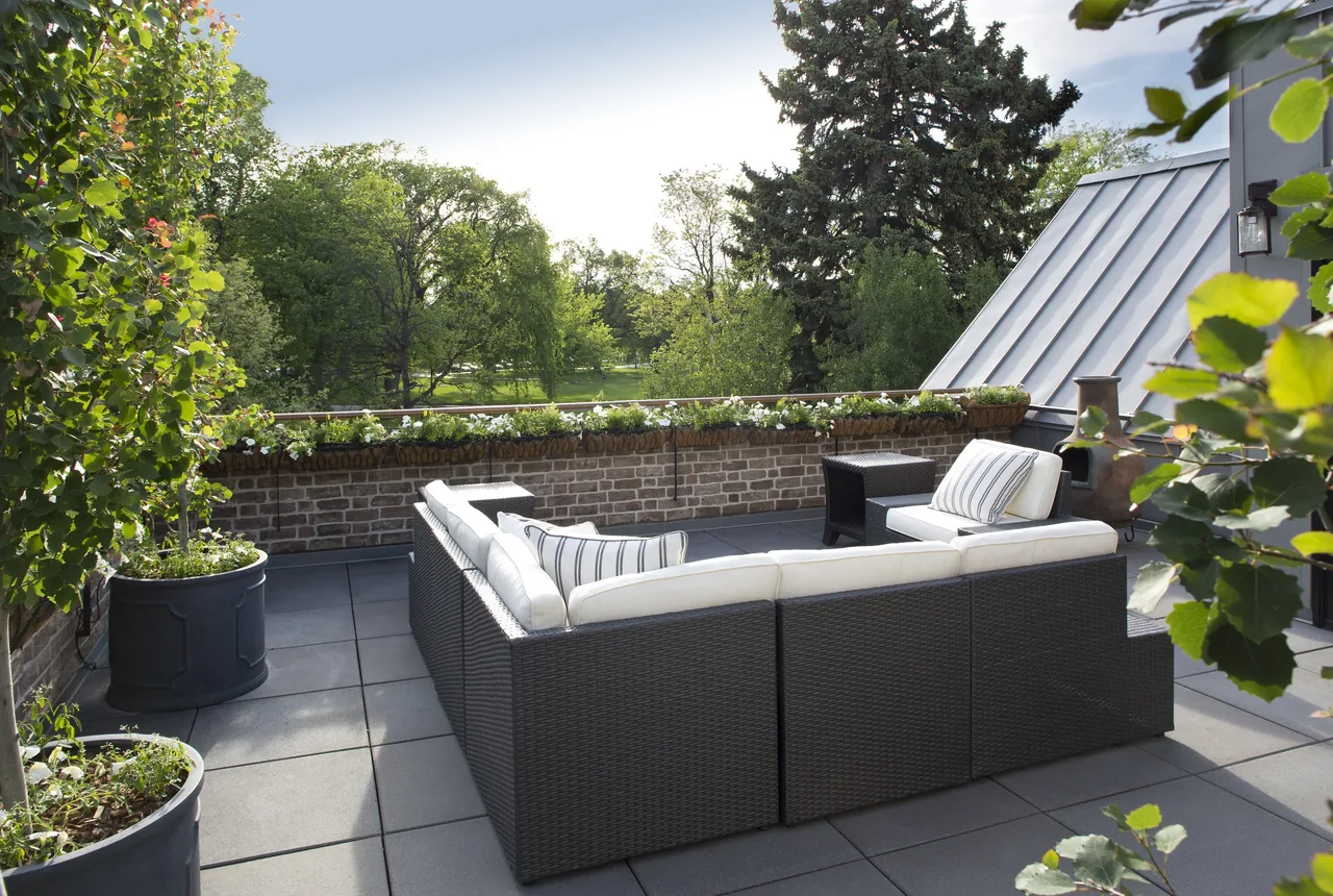 Patio area featuring dark woven outdoor sectional sofa with white cushions, surrounded by brick wall and greenery.
