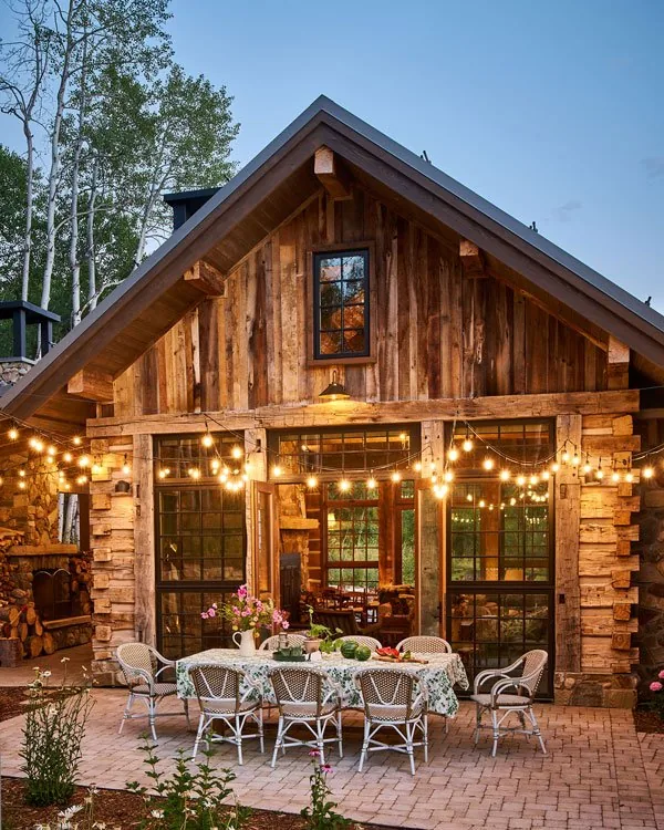 Rustic homestead patio with wood siding, stone chimney, and string lights over outdoor dining area.
