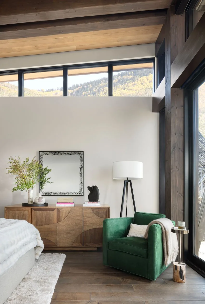 A corner of a bedroom with a green armchair, wooden sideboard, floor lamp, and high clerestory windows showing a mountain view.