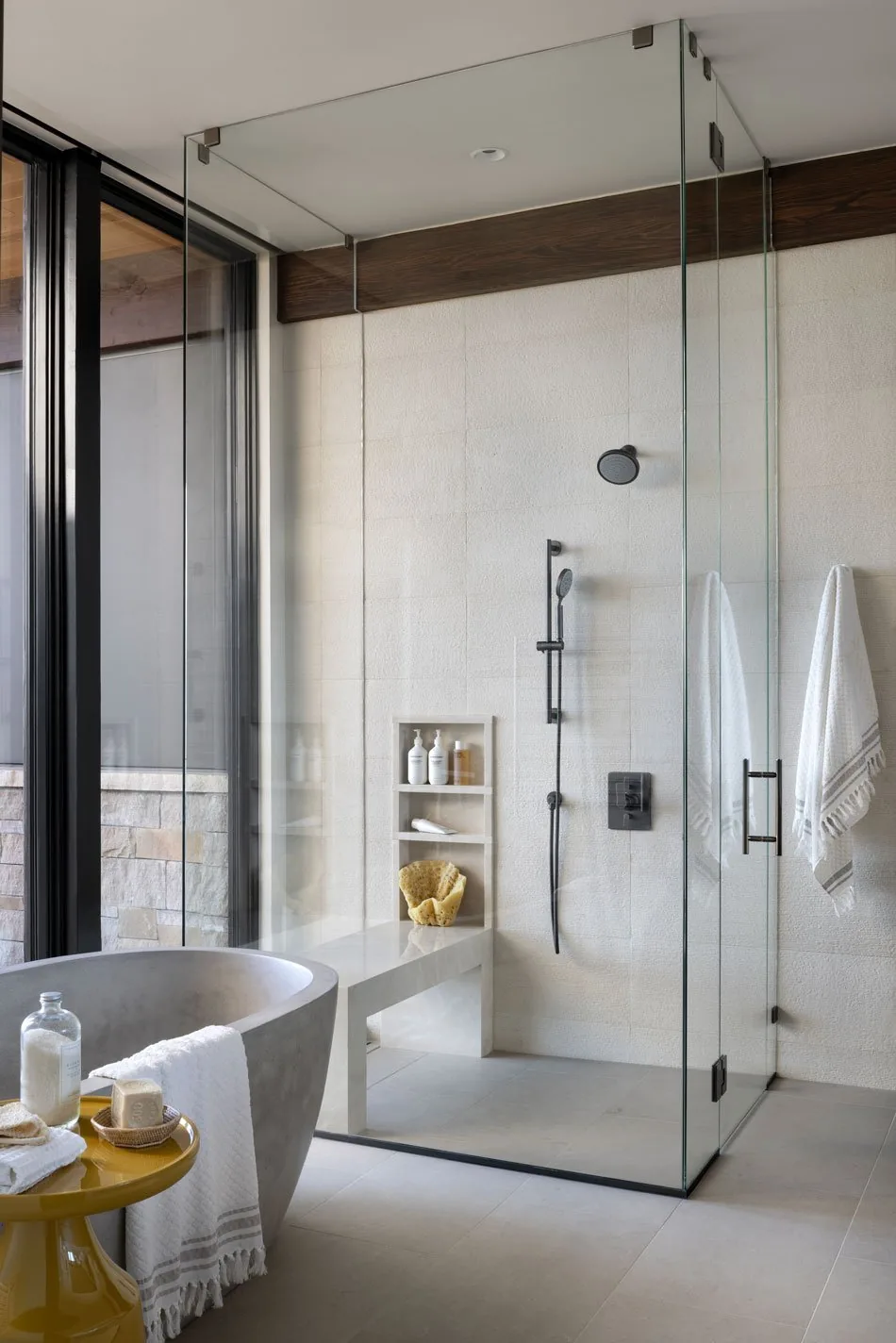 A neutral-toned bathroom with a frameless glass shower enclosure, textured wall tiles, and concrete freestanding tub.
