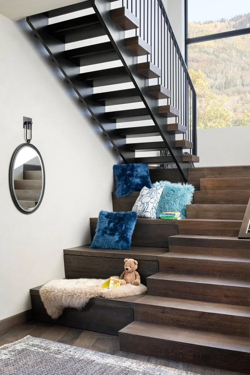 Interior view of dark wood stairs with open risers and black metal railings, featuring a sheepskin throw, pillows, and a round wall mirror.