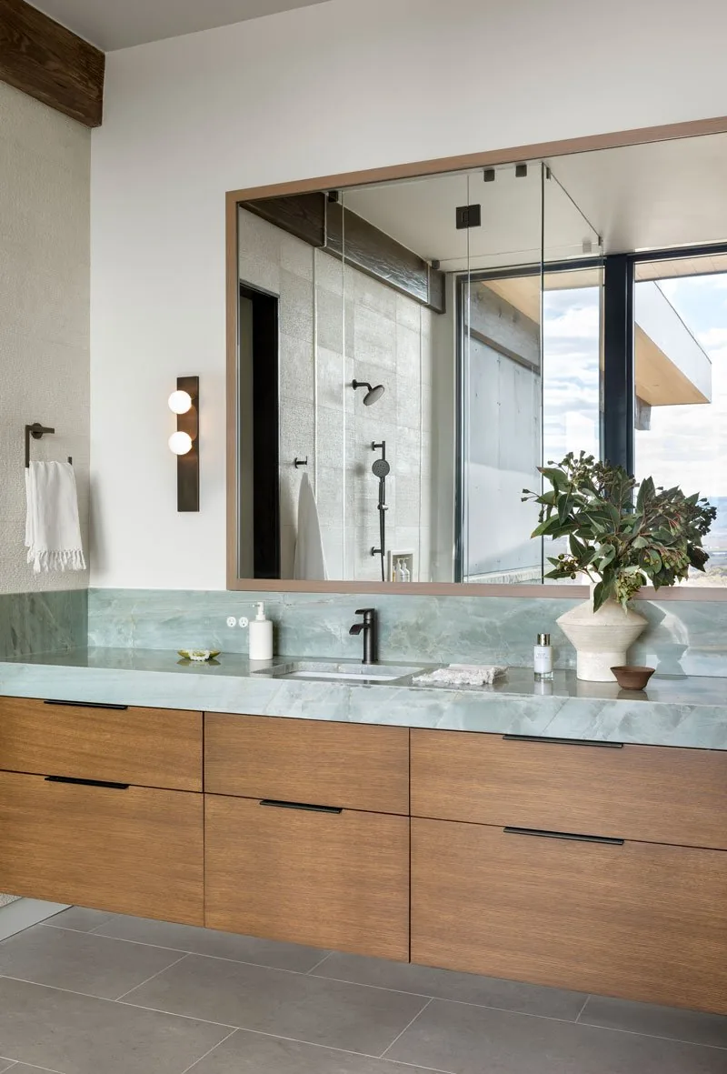 Bathroom vanity area with floating wooden cabinets, green stone countertop, black faucet, and large mirror reflecting a glass-enclosed shower.