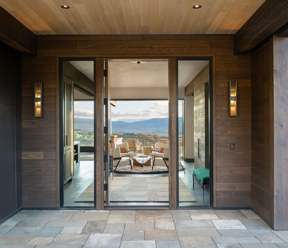 Wooden framed glass doors open into a sitting area featuring modern chairs and expansive mountain views.