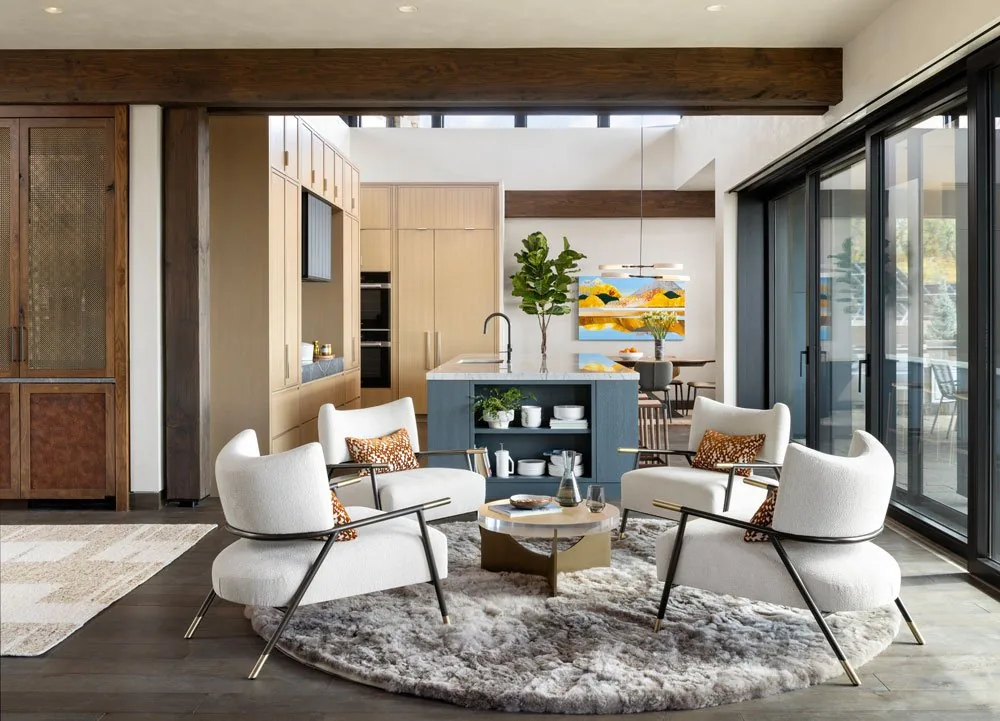 Open living and kitchen area with armchairs around coffee table and kitchen island in background under exposed wood beams.