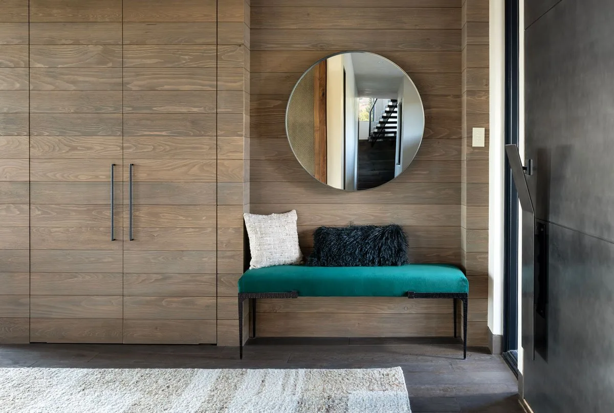 Entry with wood cabinetry, teal bench seating, circular mirror, and dark pivot door.