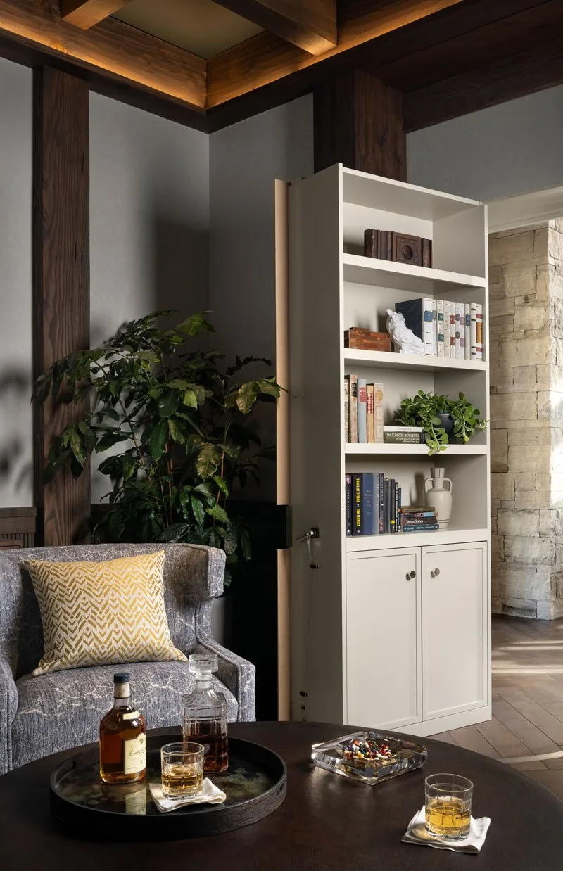 A living room corner with a gray armchair, white bookshelf, and stone wall opening to another space.