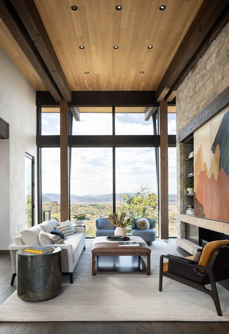 Interior living room featuring floor-to-ceiling glazing, stone fireplace, wood ceiling with exposed beams, and mixed seating arrangement.
