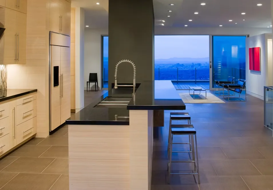 Interior view of a modern kitchen and living area with large sliding glass doors opening to a balcony overlooking a cityscape at dusk.