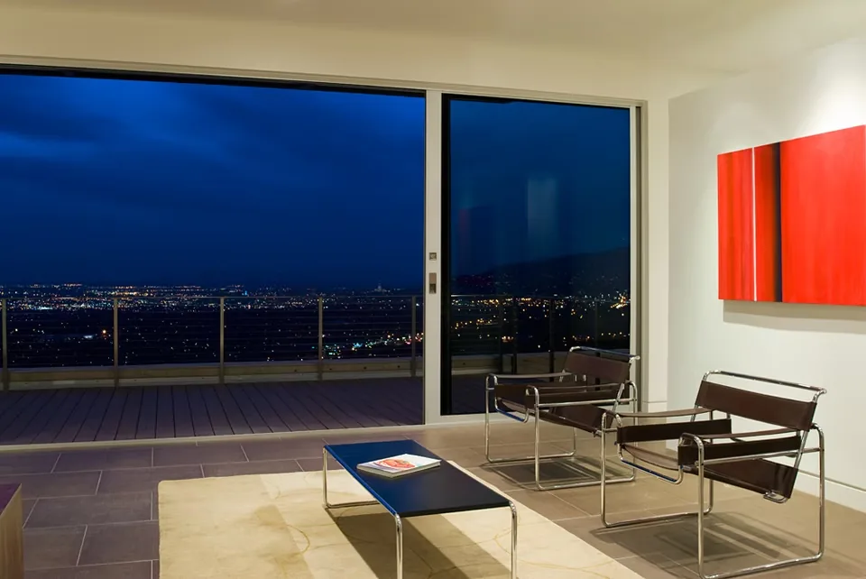 Modern living room with metal-framed chairs and glass coffee table opening to a balcony through sliding glass doors at night.