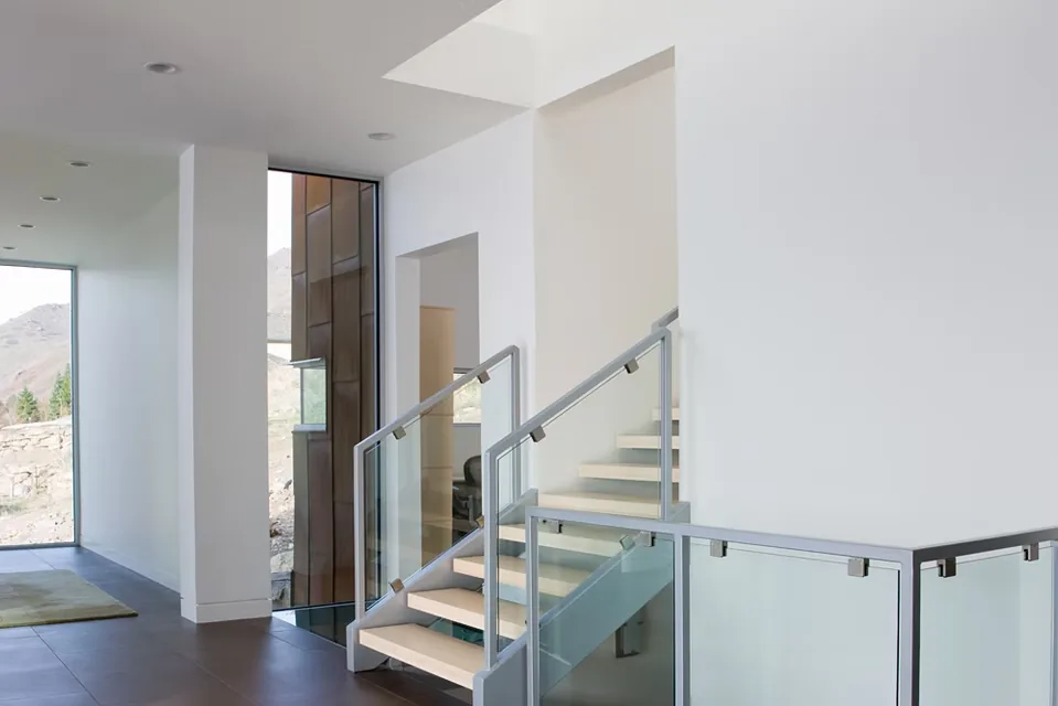 Modern staircase in a residential interior with glass and metal railing next to floor-to-ceiling windows.
