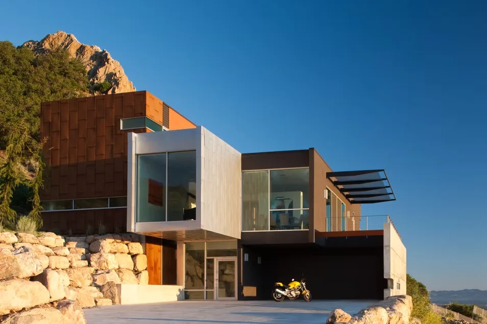 Exterior front view of a modern cantilevered house featuring corten steel, concrete, and floor-to-ceiling glass windows on a rocky site.