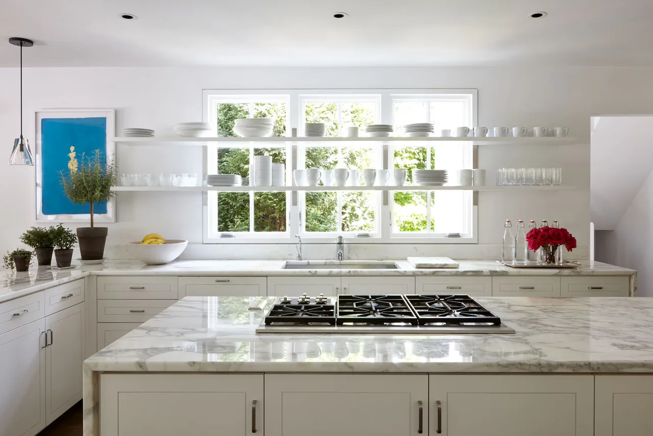 Kitchen with marble countertops, gas cooktop island, and open shelves against window.