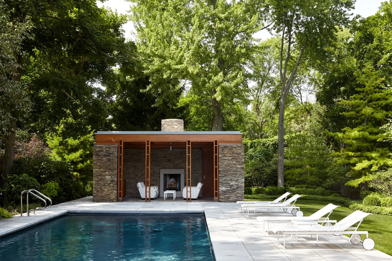 Rectangular outdoor pool with stone pool house featuring a fireplace and adjacent lounge chairs in a wooded setting.