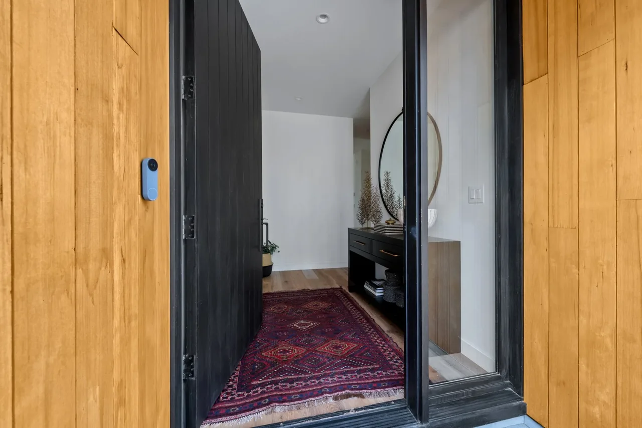 Entryway with vertical wood siding and a pivot door opening to a hallway with a rug and console table topped by a round mirror.
