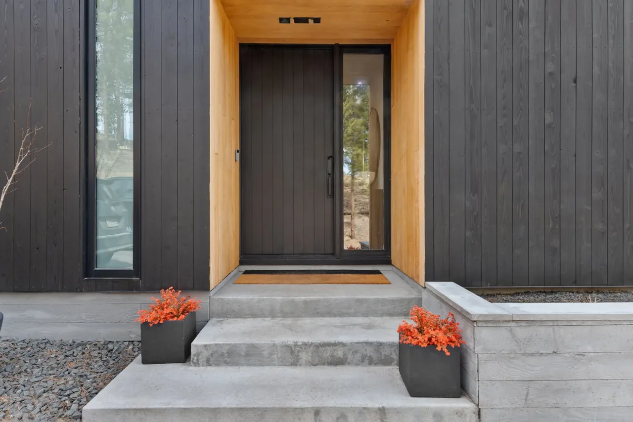 The entryway features a black vertical wood pivot door, framed by light wood paneling and concrete steps with potted plants.
