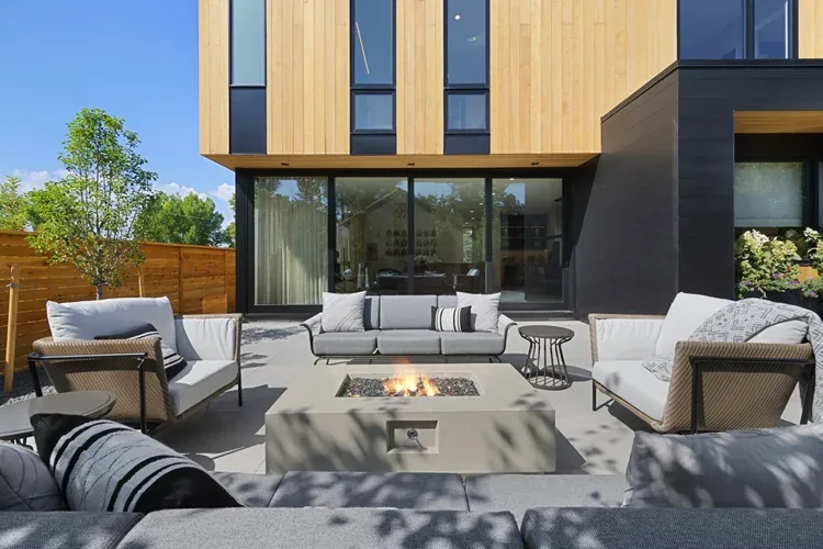Outdoor patio seating arranged around a central fire pit with floor-to-ceiling glass windows and wood paneling on the building exterior.