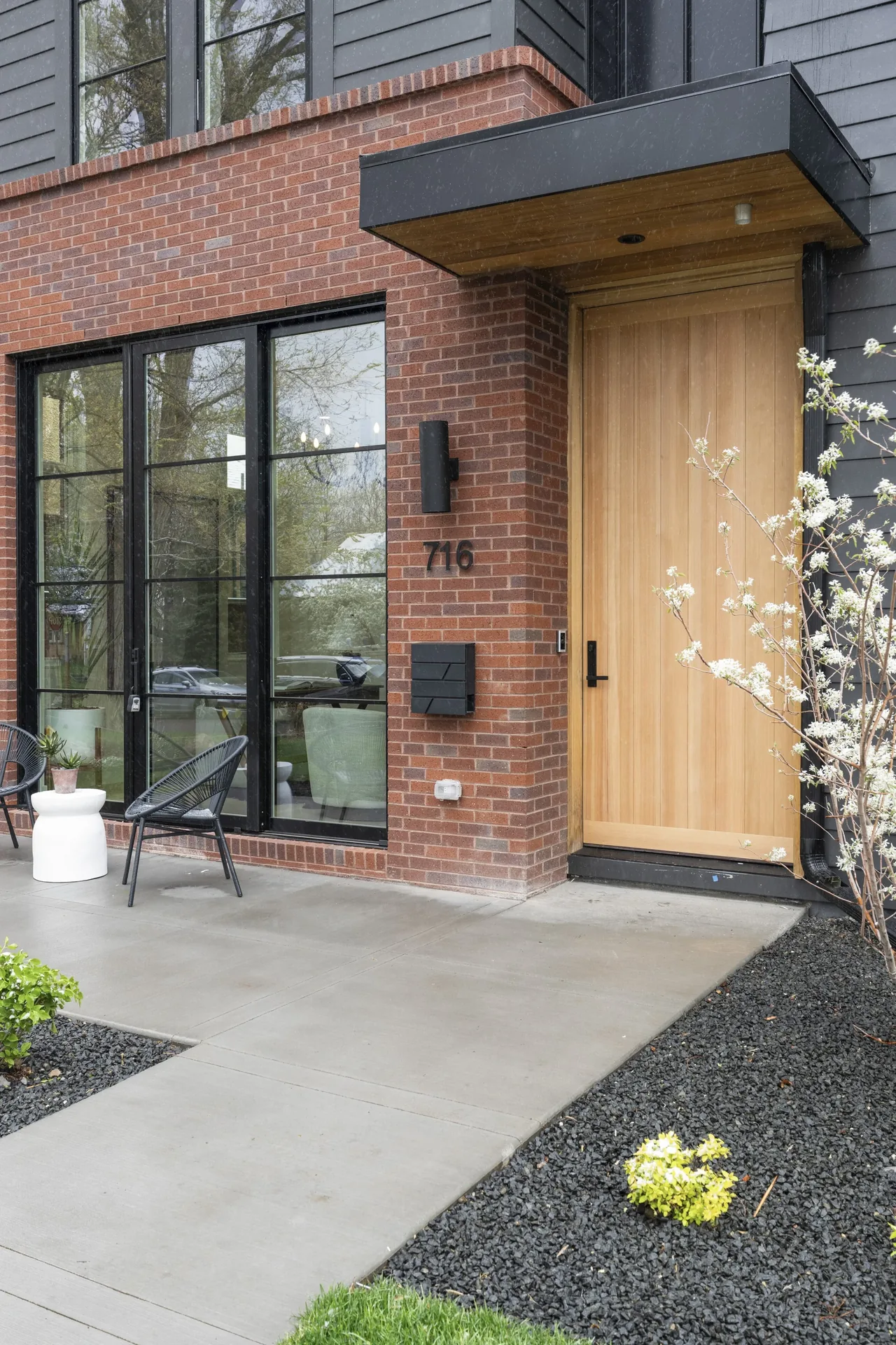 Front entrance with a light wood door, black metal-framed windows, and brick wall with concrete walkway and planted landscaping.