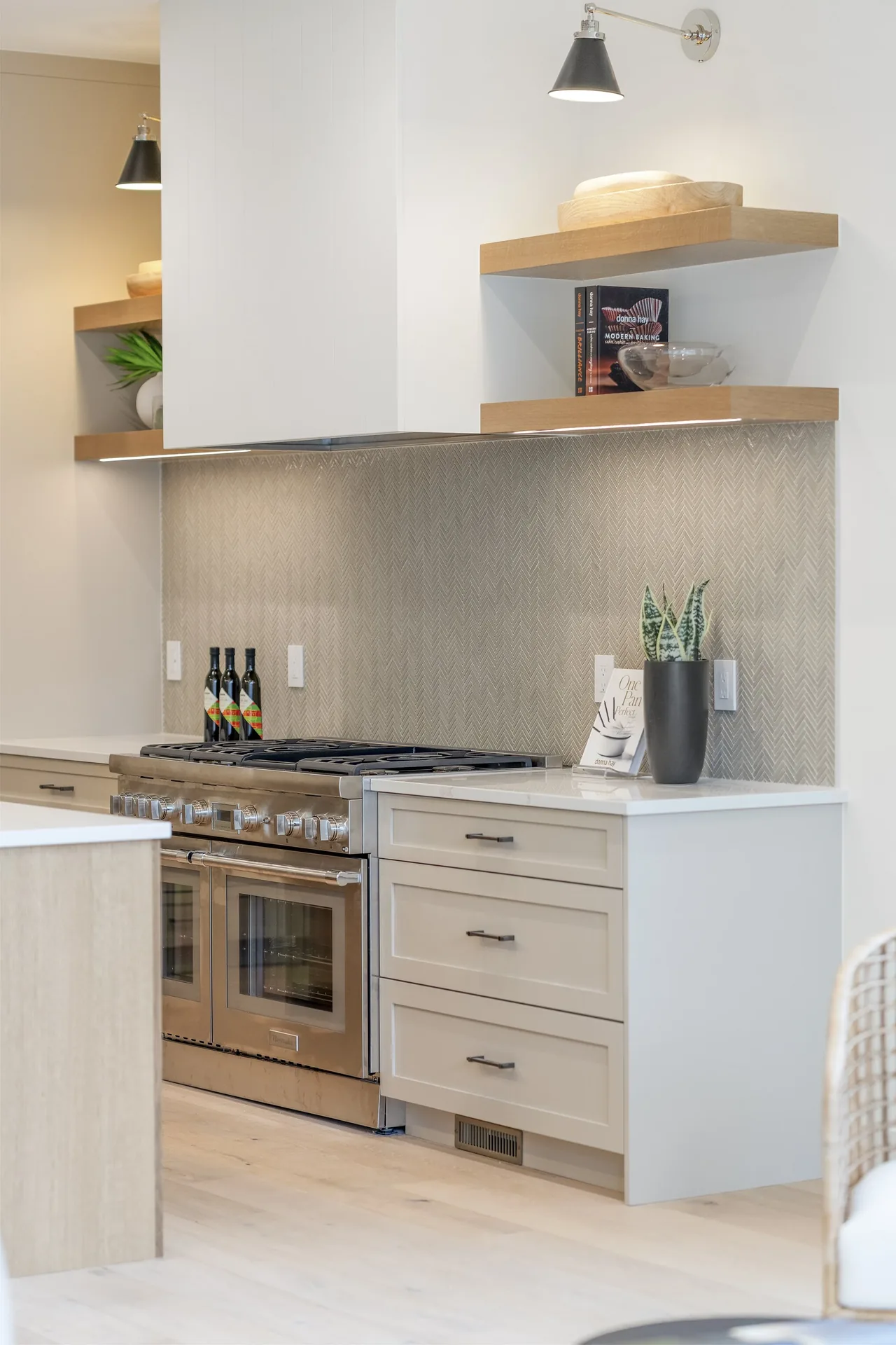 Kitchen with stainless steel gas range, white cabinetry, wood floating shelves, and a herringbone pattern tile backsplash.