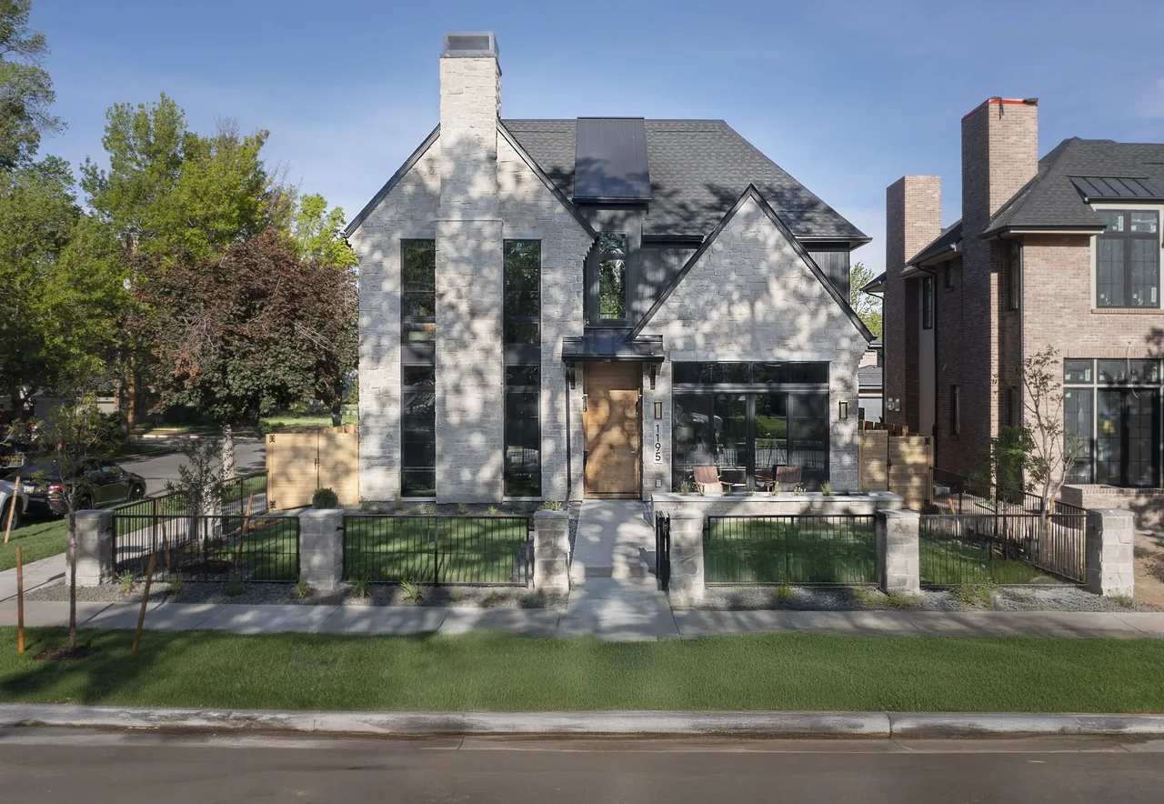 Front elevation of a two-story stone house with gable roof, vertical windows, and fenced front yard.