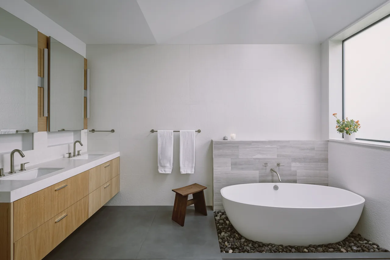 Bathroom with freestanding tub on pebble floor and double vanity with wood cabinetry under rectangular mirrors.