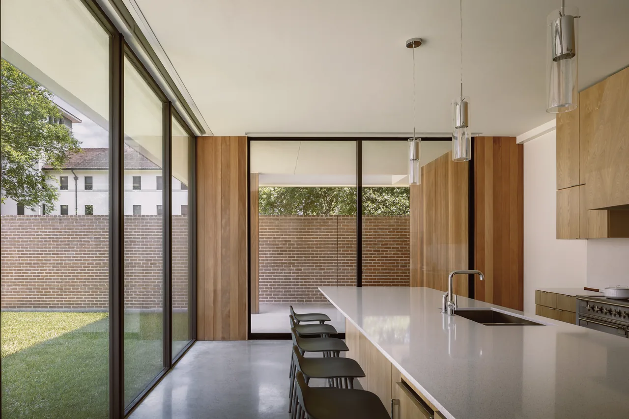 Kitchen in Rice Magister House with wood cabinets, a white island, pendant lights, and large sliding glass doors facing a brick privacy wall.