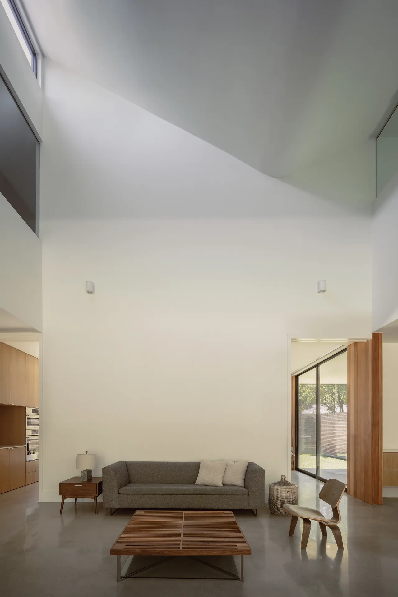 Living room interior with high ceiling, natural wood furniture, and a large window opening to a garden.