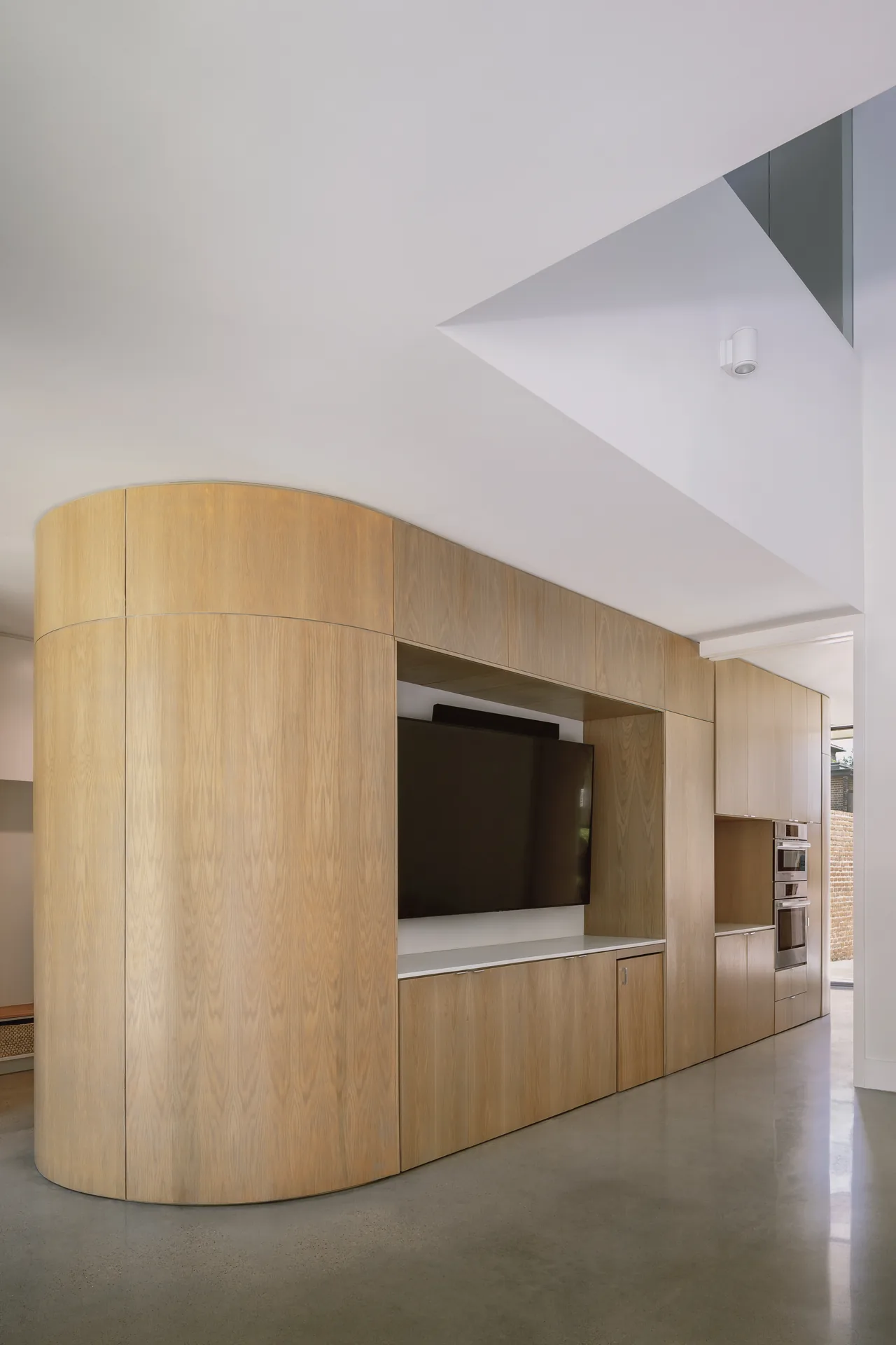 Curved wood-paneled cabinetry integrates a mounted TV and kitchen ovens in a modern open-plan interior.