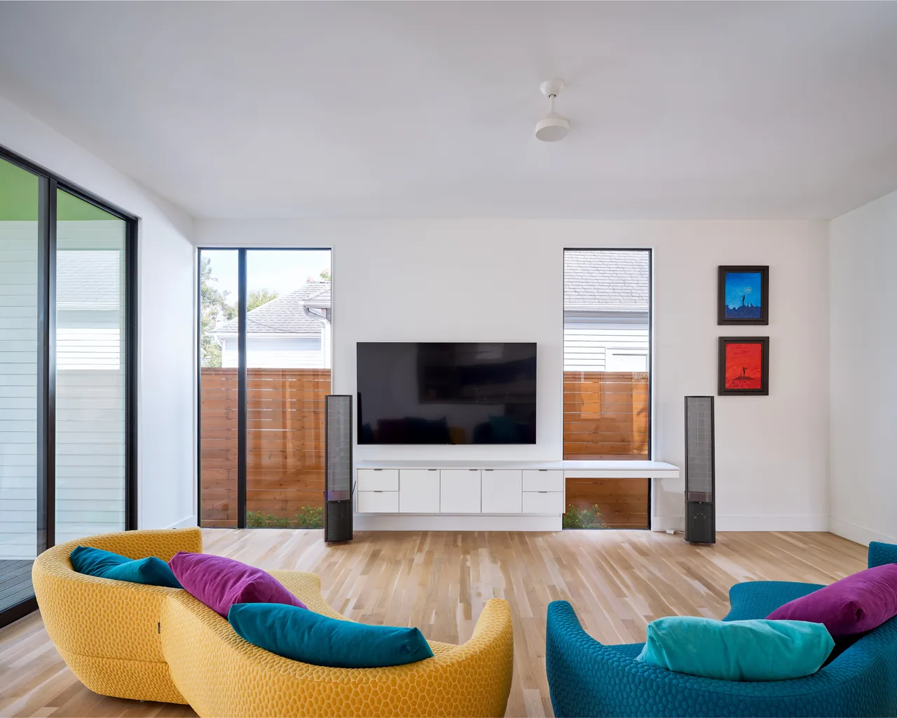 Living room features curved yellow and blue chairs with cushions, mounted TV between tall windows, and two colorful paintings on a white wall.