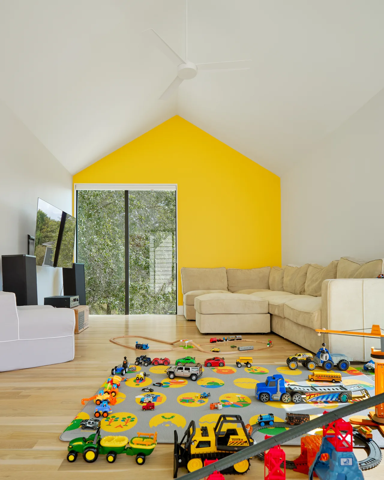 The Allston Residence living room features a vaulted ceiling, yellow accent wall, and hardwood floors, furnished with a beige sectional and children's play area.