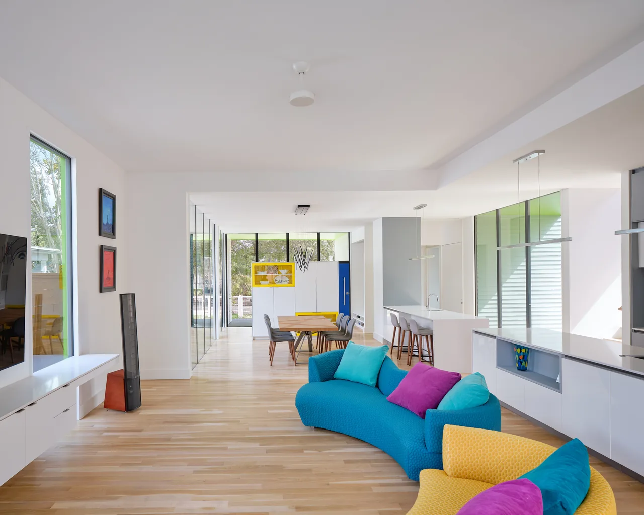 Open-plan living and dining area with curved sofas in blue and yellow, colorful pillows, and large windows around the space.