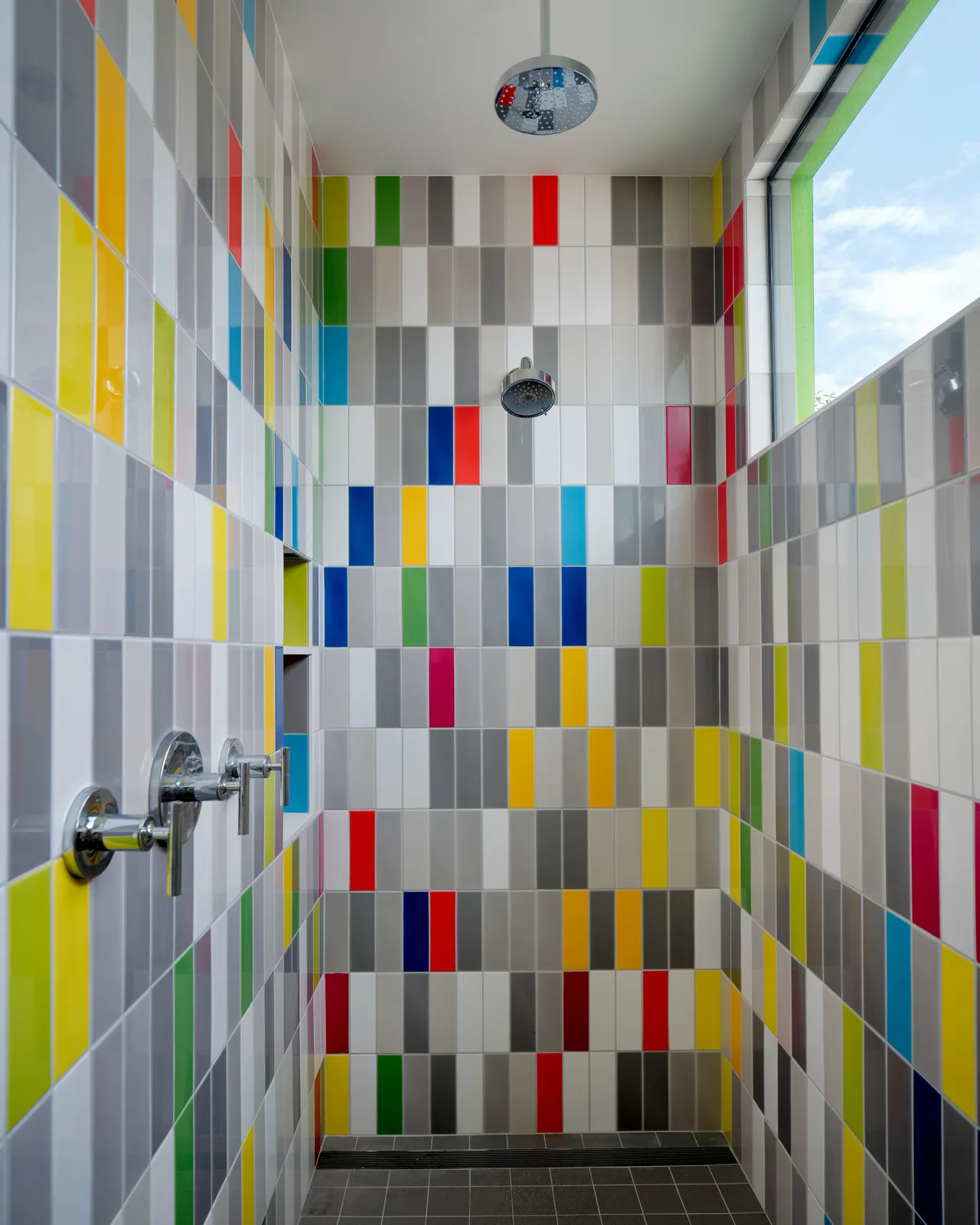 Dual shower heads in a colorful tiled shower with a rain shower head overhead and a large window.