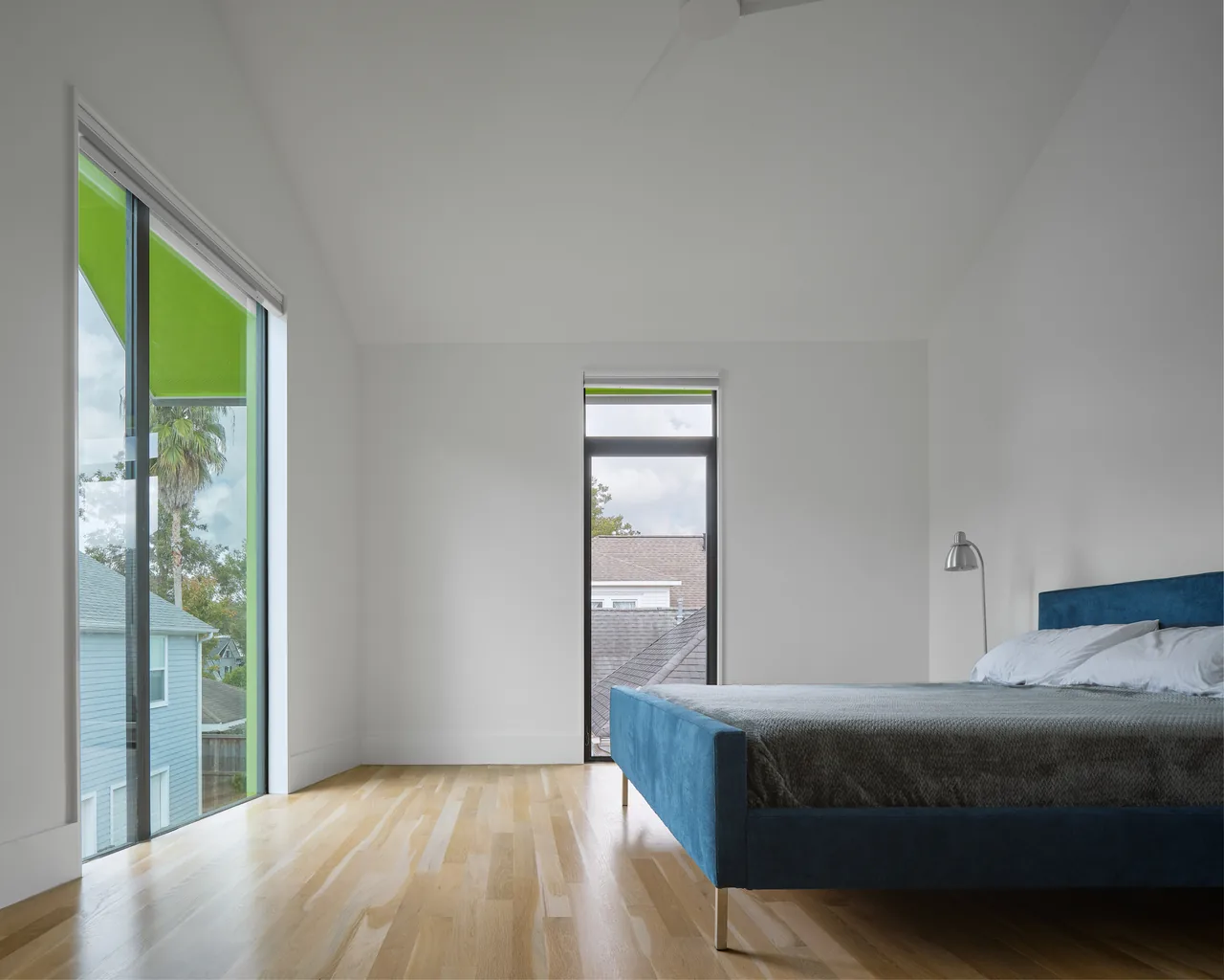The Allston Residence bedroom features clean lines, large windows with green trim, and a blue upholstered bed on light hardwood flooring.