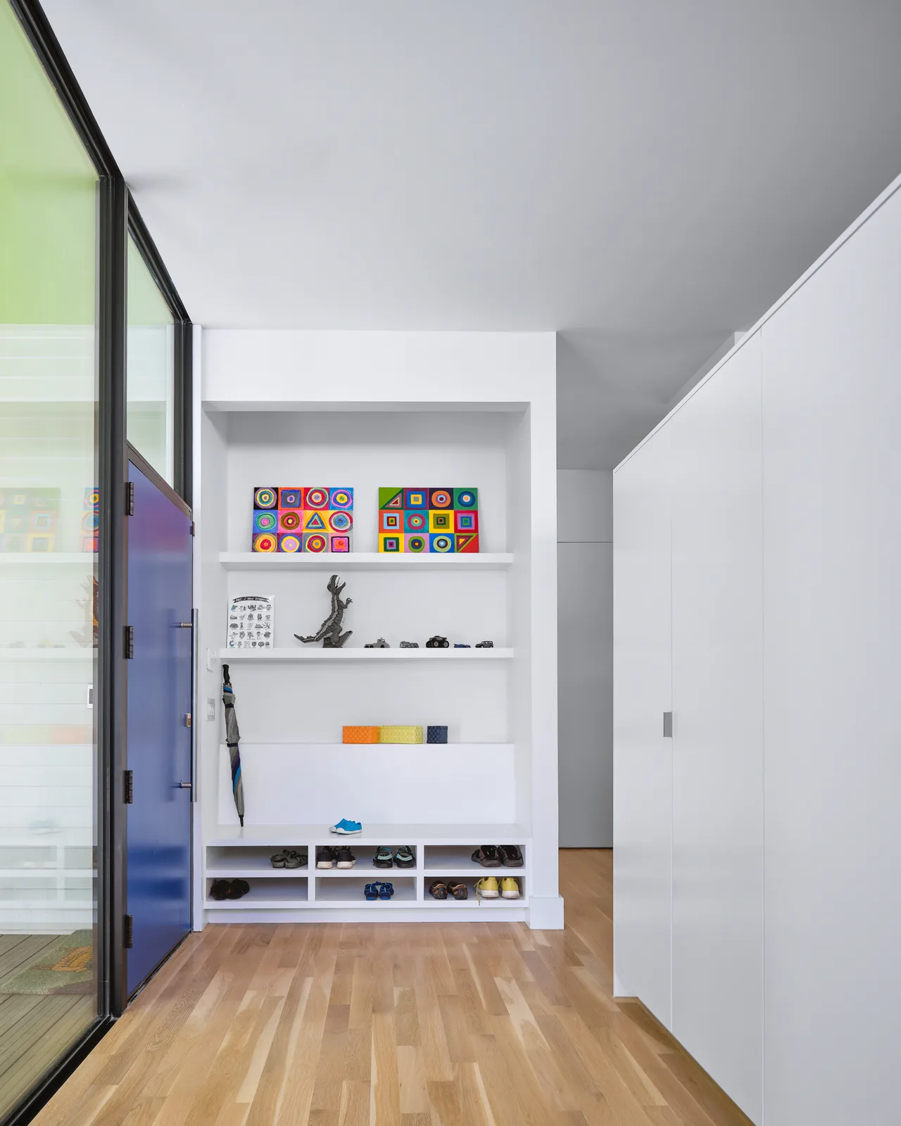 Entry area of the Allston Residence features built-in shelving with art and shoe storage, a blue front door, and bright green window trim.