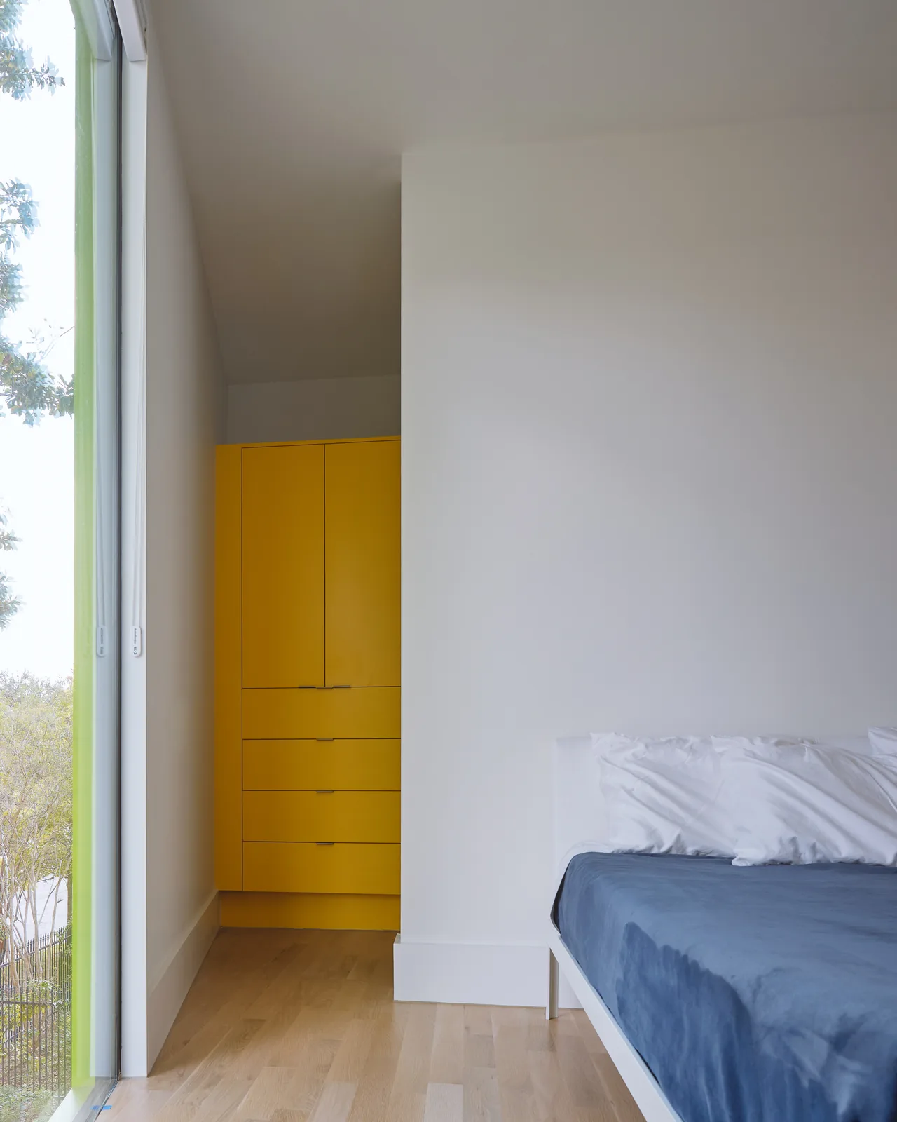 Corner view of a minimalist bedroom featuring a yellow built-in wardrobe and a bed with blue bedding at Allston Residence.