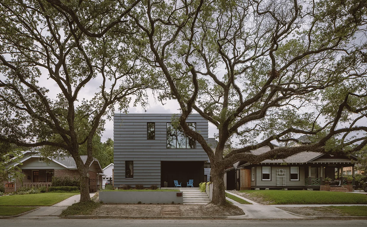 Modern two-story house with horizontal gray cladding and front porch, bordered by large trees and adjacent to traditional single-story homes.
