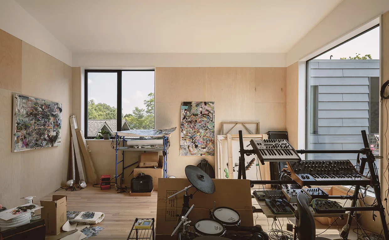 A room features light wood panel walls and floor, artistic work, and audio equipment near large rectangular windows.